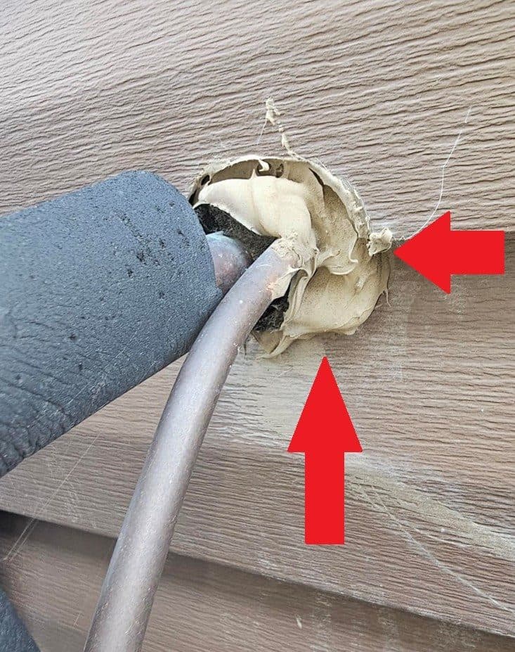 Refrigerant lines entering siding through a rough hole packed with cracked foam and caulking.
