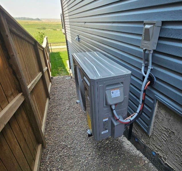An outdoor unit on a wall bracket in a gravel side yard, with its wiring and lines running straight and fastened to the wall.