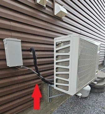 A disconnect box and outdoor unit on brown siding, with the wiring and lines between them left unsupported, marked with a red arrow.